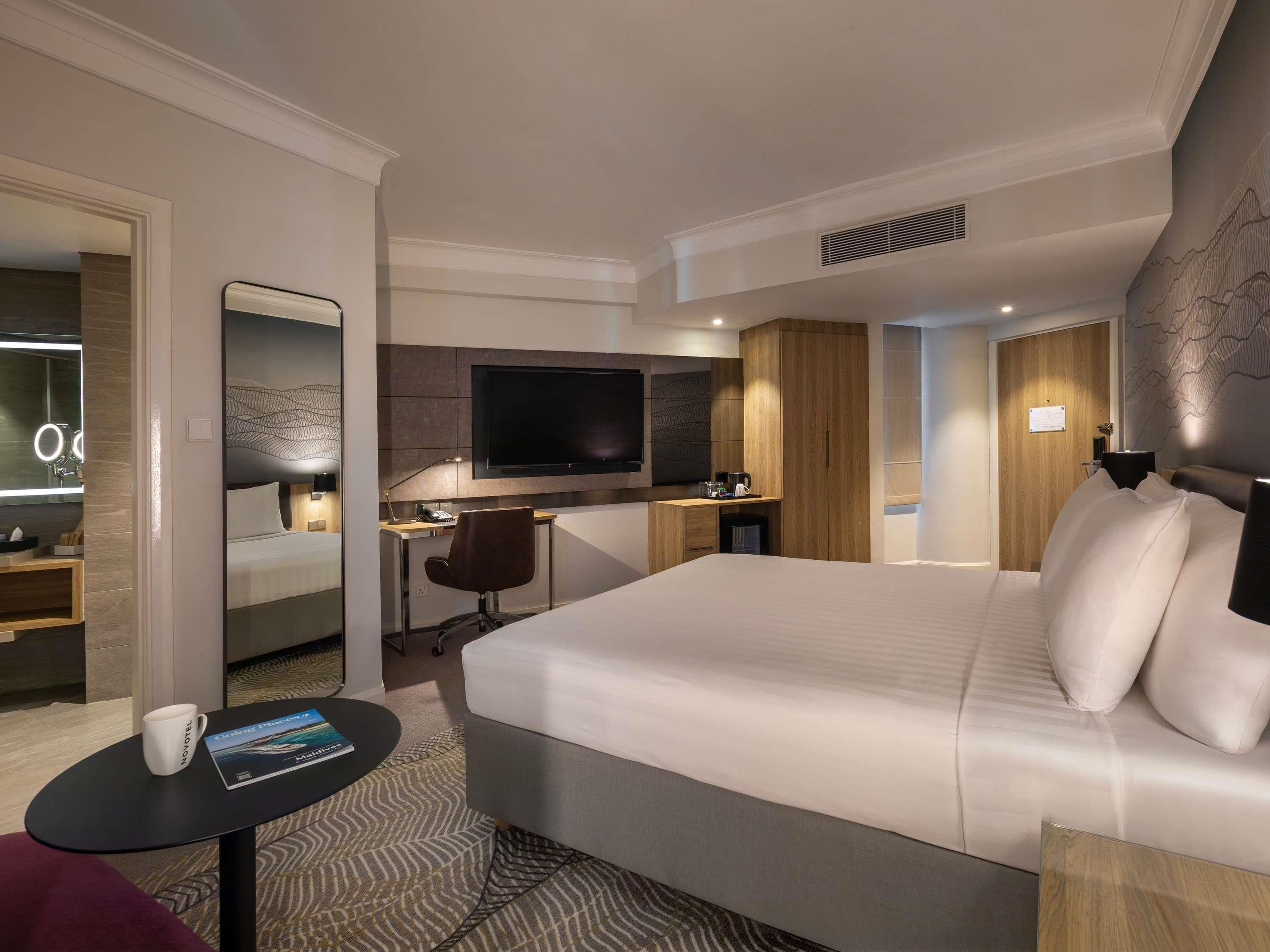 A modern hotel room with a large bed, a desk with a computer, a TV, and a bathroom with a mirror.