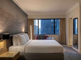 A hotel room with a large bed, a small table, a chair, a wall art piece, and a city view through the window.