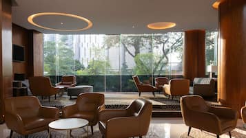 A modern hotel lobby with leather armchairs, a round table, and large windows offering a view of trees and buildings.