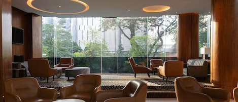 Un moderno vestíbulo de hotel con sillones de cuero, una mesa redonda y ventanas amplias con vistas a árboles y edificios.