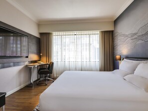 Una habitación de hotel con una cama grande, un escritorio con silla, televisión y ventana con cortinas.
