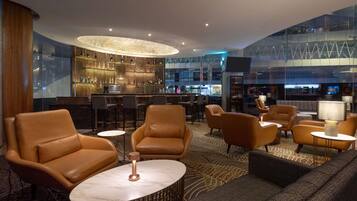 A modern lounge area with leather sofas, a bar with a glass wall, and a high ceiling with recessed lighting.