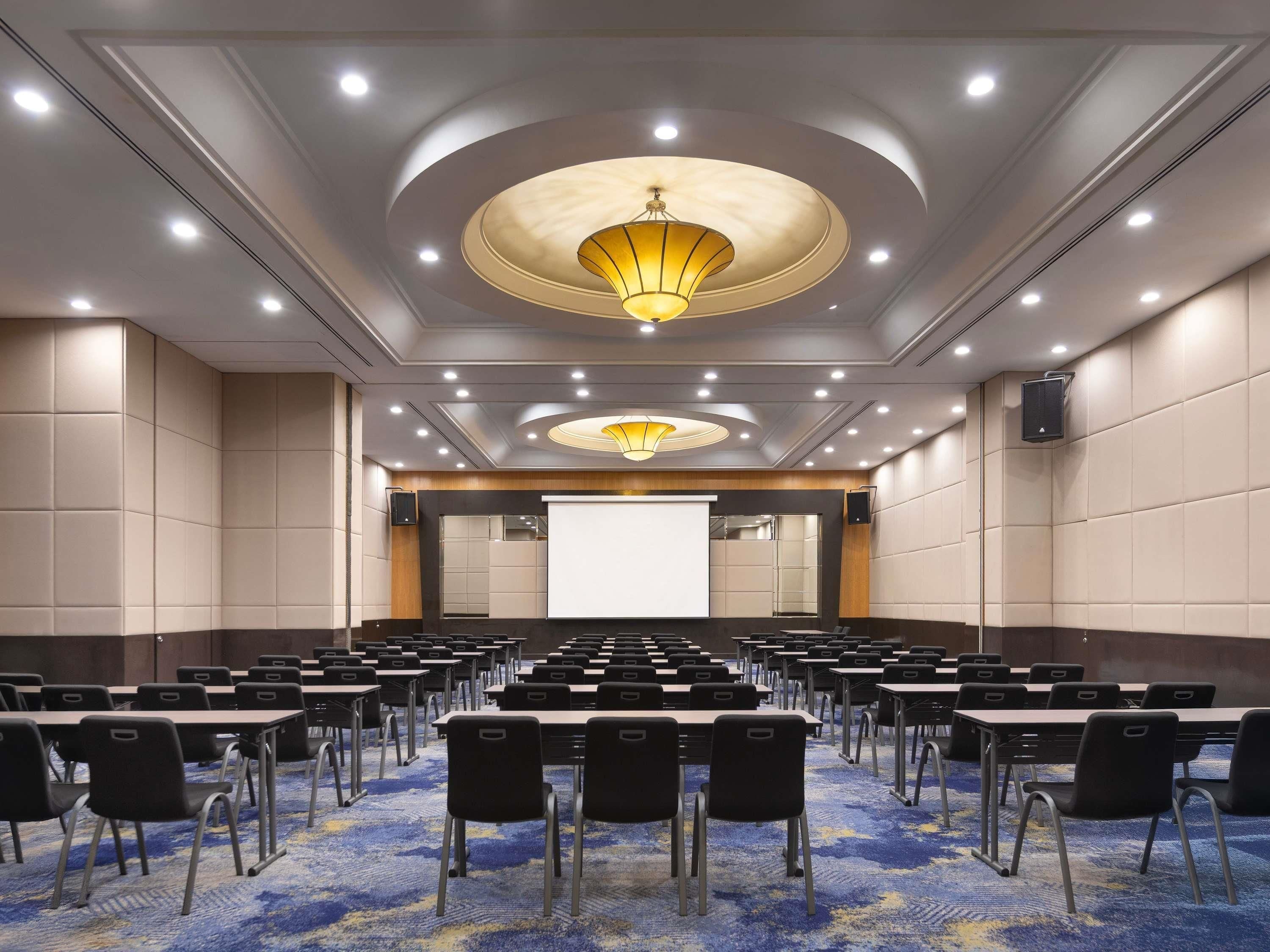 A conference room with rows of tables and chairs, a large screen, and a chandelier.