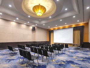 Un salón de conferencias con filas de sillas, una pantalla para proyector y una lámpara grande en el techo.