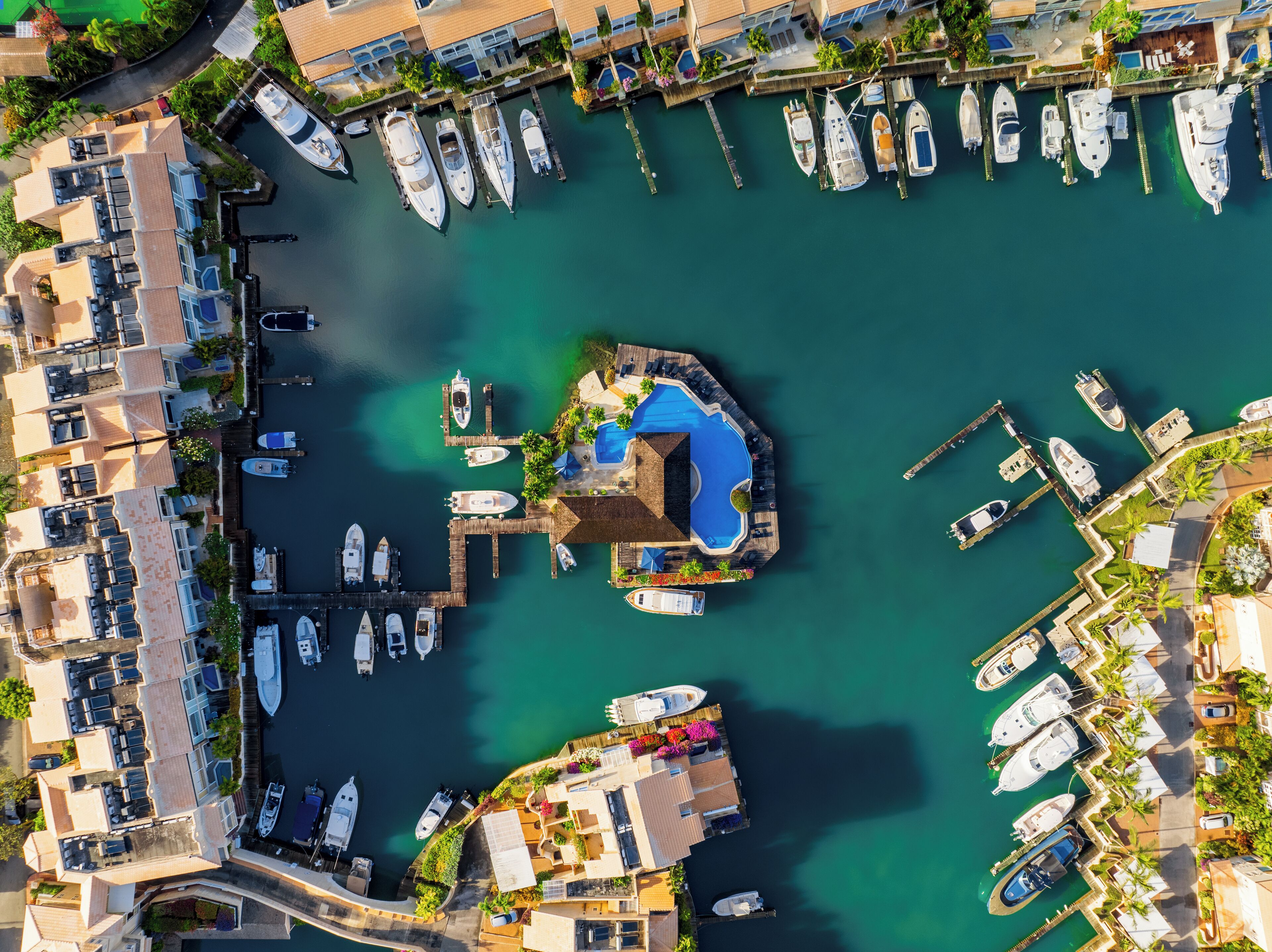 An aerial view of a marina with multiple boats docked, a central swimming area, and surrounding residential buildings.