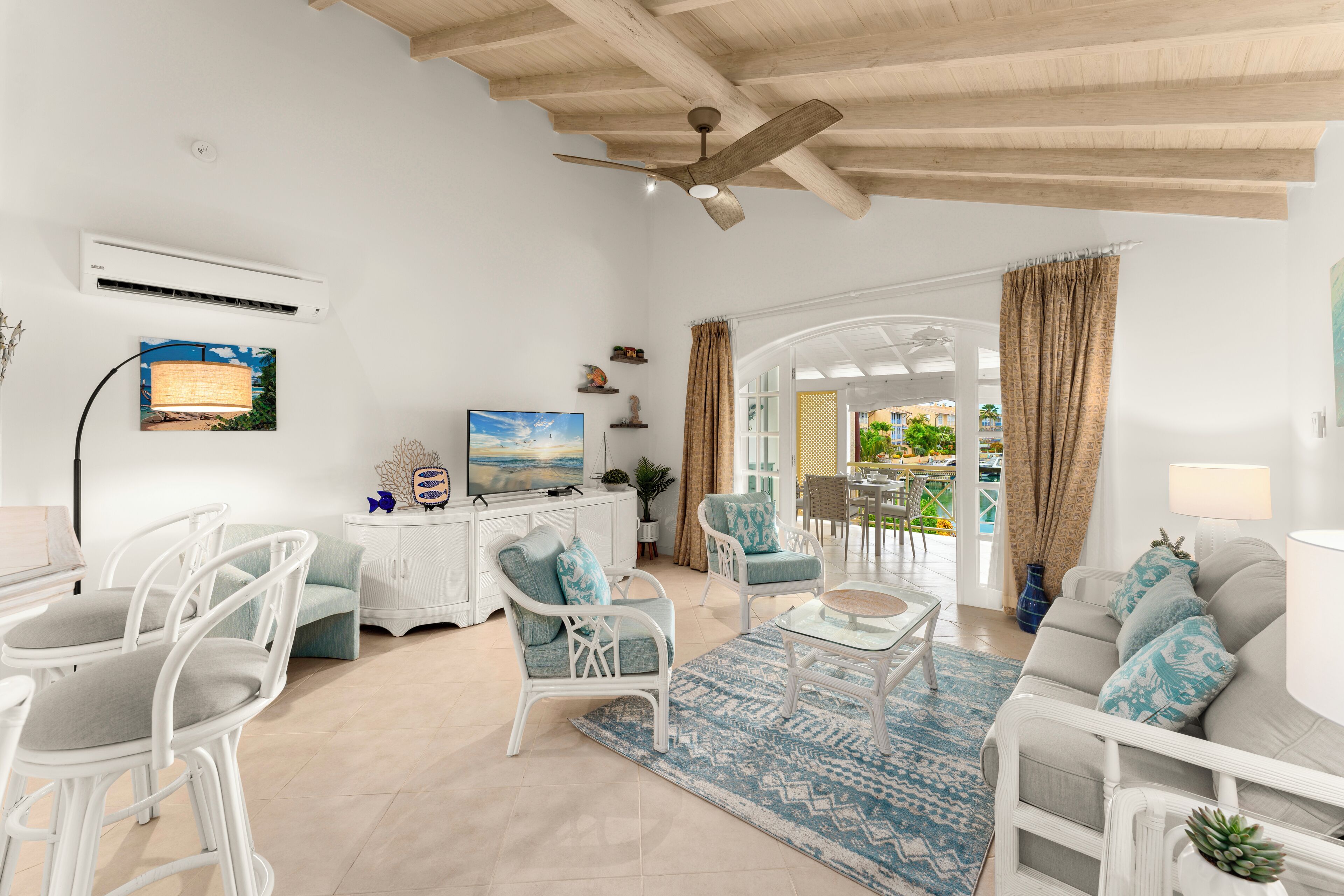 Un salón amplio con muebles blancos, un ventilador de techo y vistas a un área exterior por una puerta abierta.