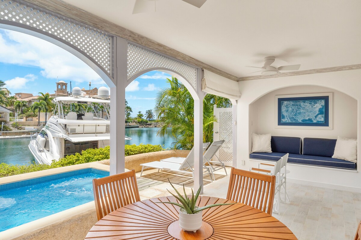 A poolside area with a table, chairs, a sofa, and a view of a boat and palm trees.