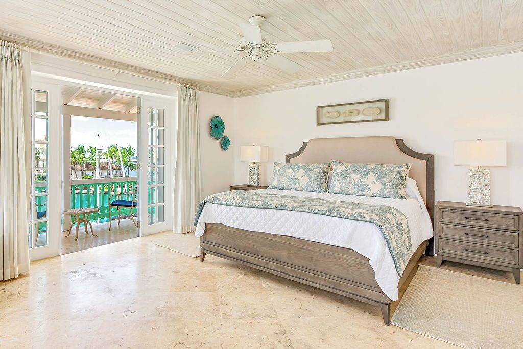 A bedroom with a large bed, a ceiling fan, a nightstand, and a view of a balcony with outdoor furniture.