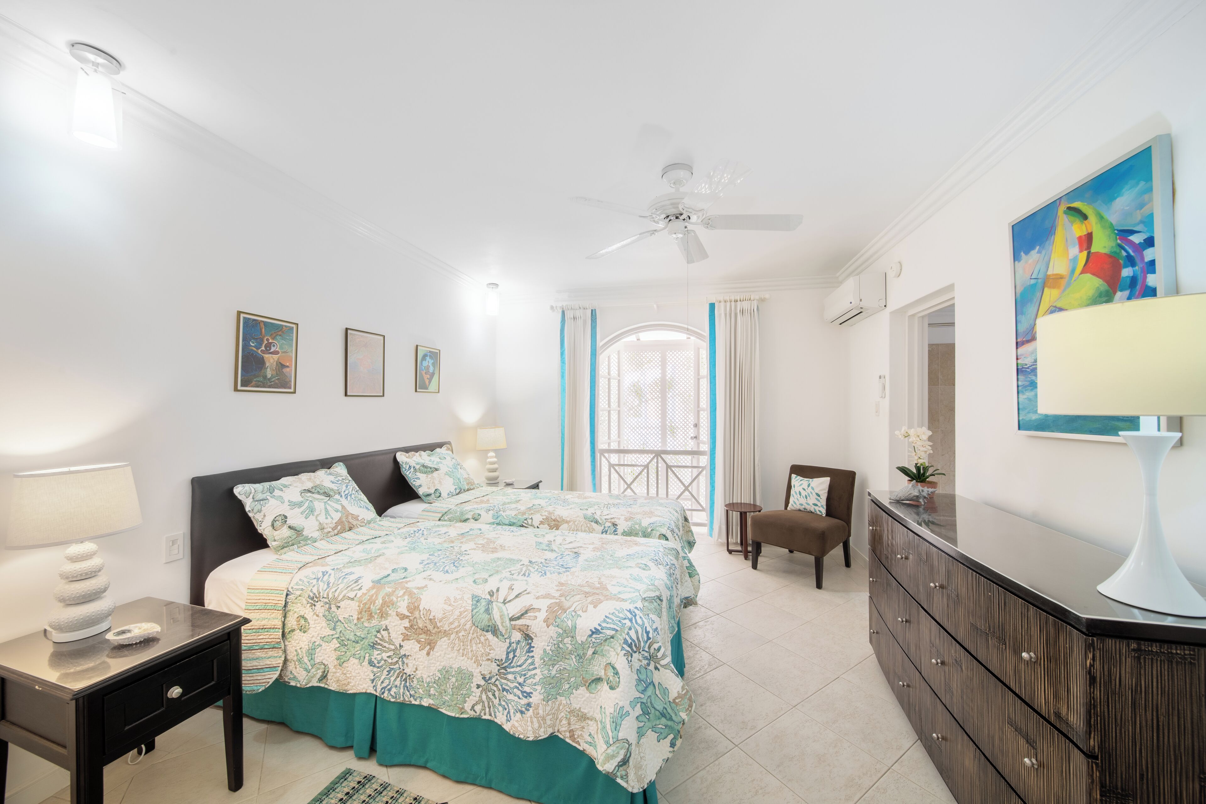 Habitación de hotel con dos camas, ventilador de techo, ventana grande con cortinas, un armario y obras de arte en las paredes.