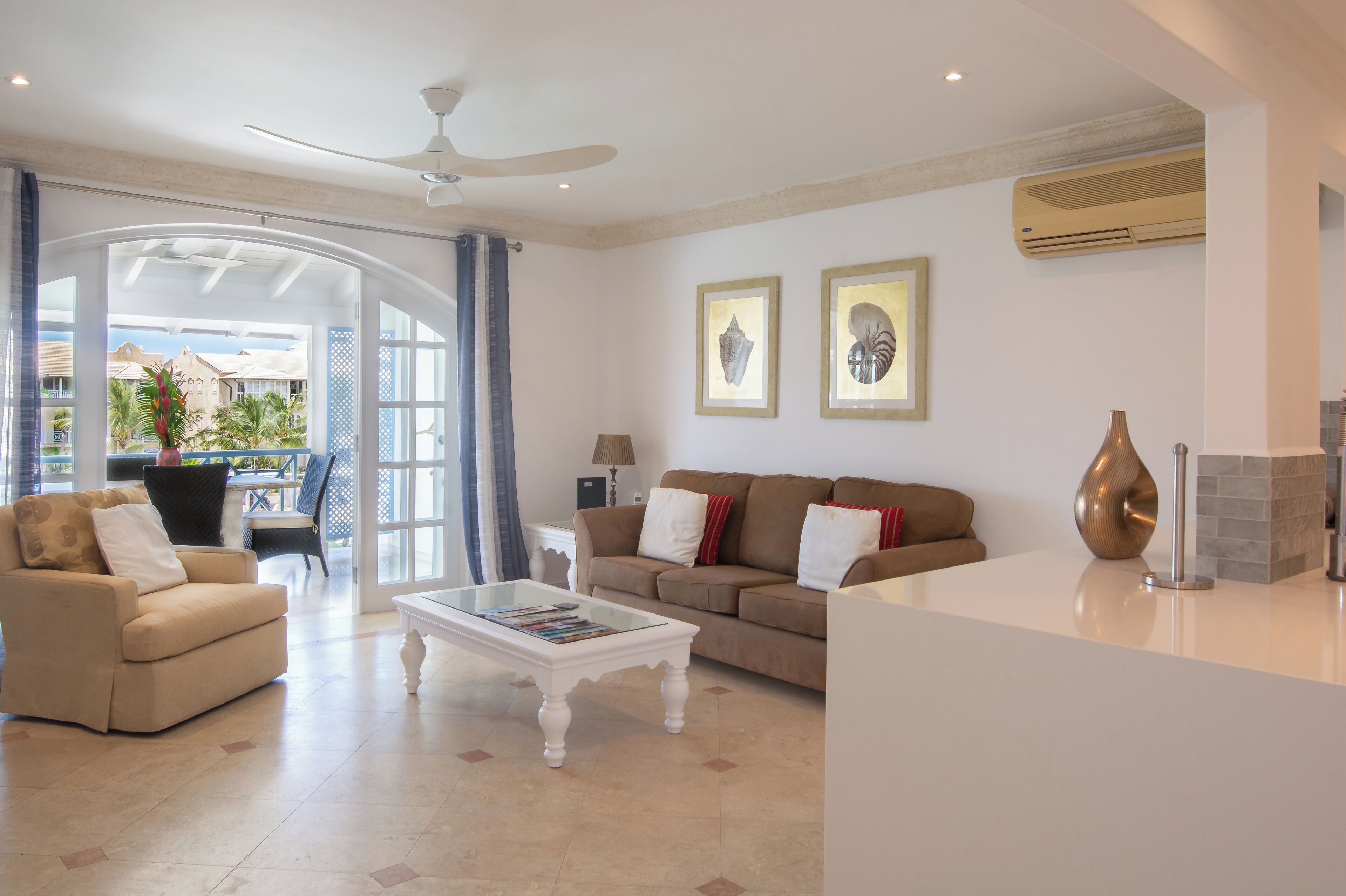 A spacious living room with a sofa, armchair, and coffee table. There is a ceiling fan, an air conditioning unit, and framed artwork on the wall.
