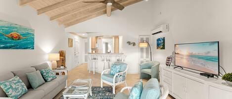 A spacious living room with a ceiling fan, a large TV, and a view of the kitchen and dining area.