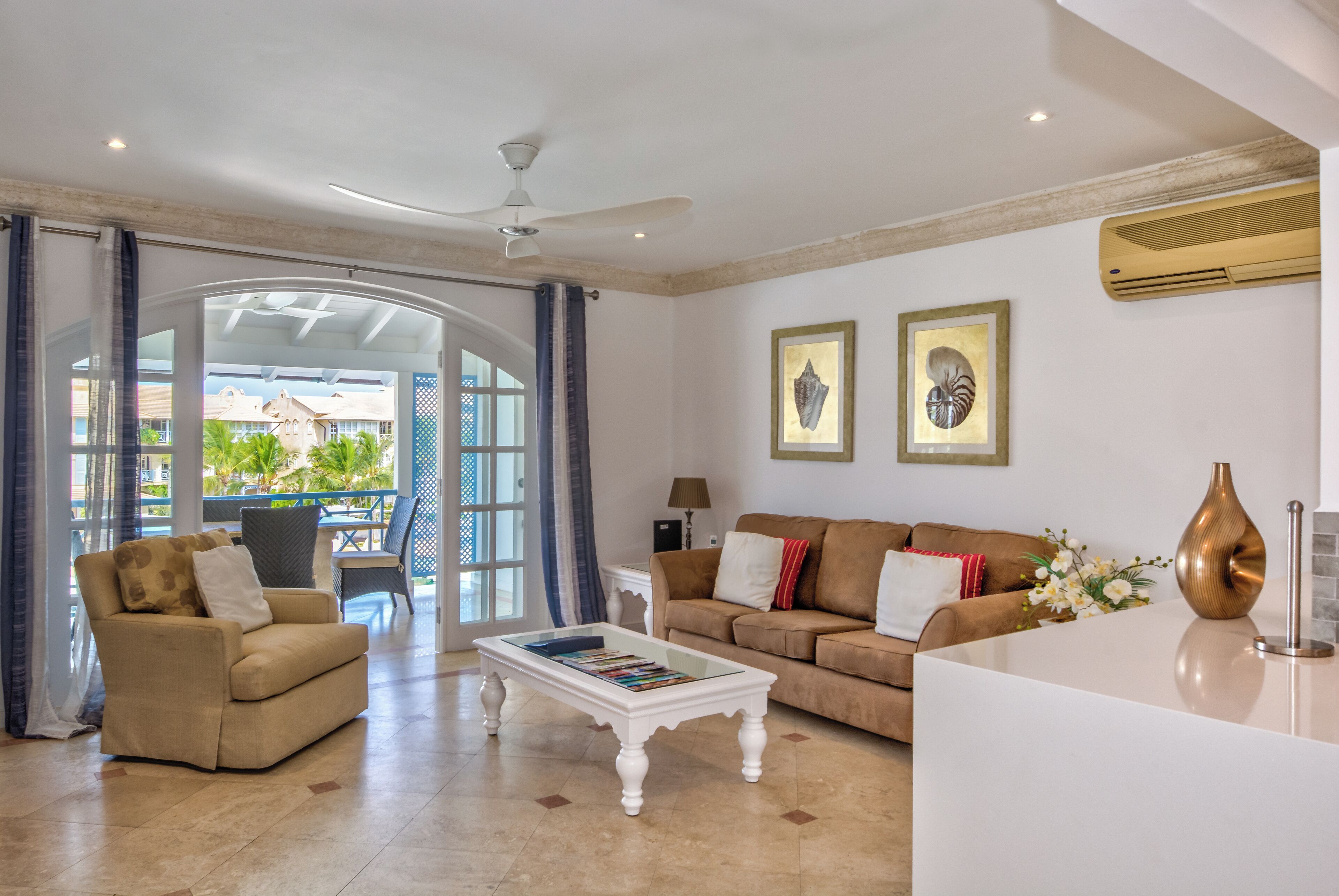 A spacious living room with a beige sofa, a white coffee table, and a brown couch. There is a glass door leading to an outdoor area with a view of palm trees.