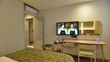 Habitación de hotel con cama, escritorio, silla, televisor mostrando dos personas y un pequeño cesto de basura.