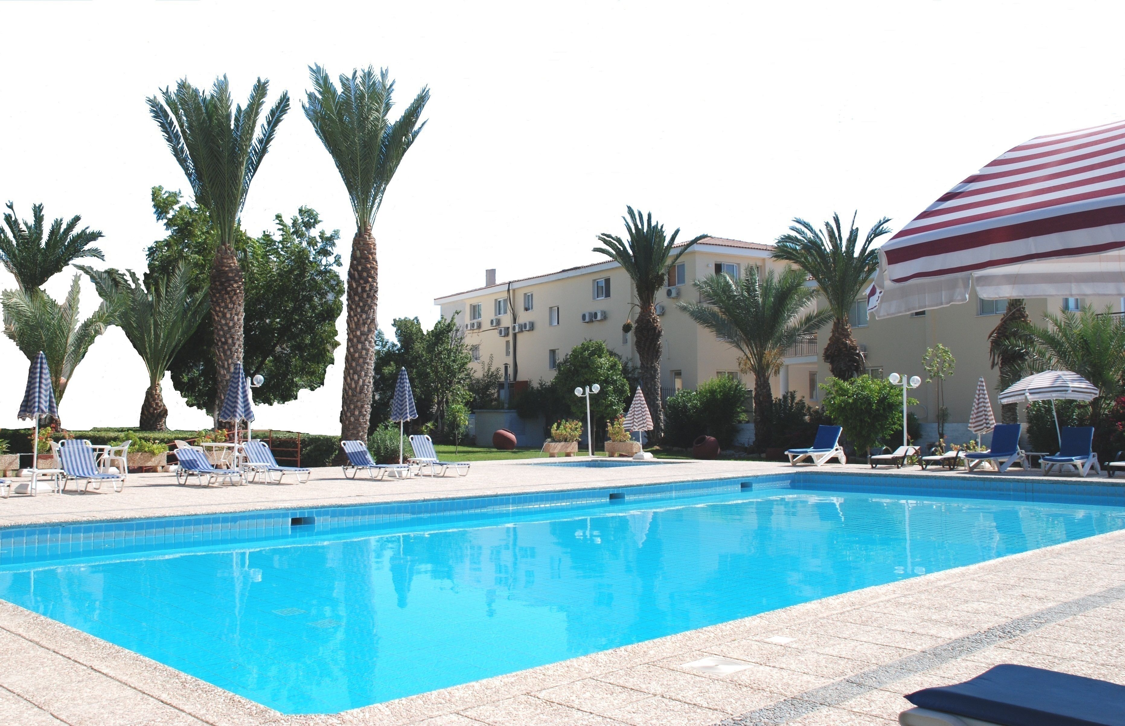 A swimming pool with lounge chairs and umbrellas, surrounded by palm trees and a building with balconies.
