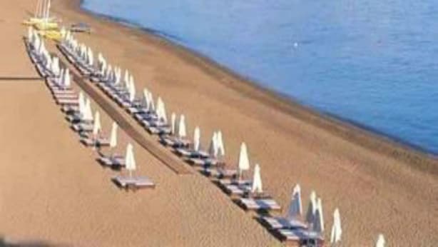 A beach with rows of sun loungers and umbrellas, a clear sky, and mountains in the background. - Marion (Polis)