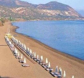 A beach with rows of sun loungers and umbrellas, a clear sky, and mountains in the background. - Marion (Polis)