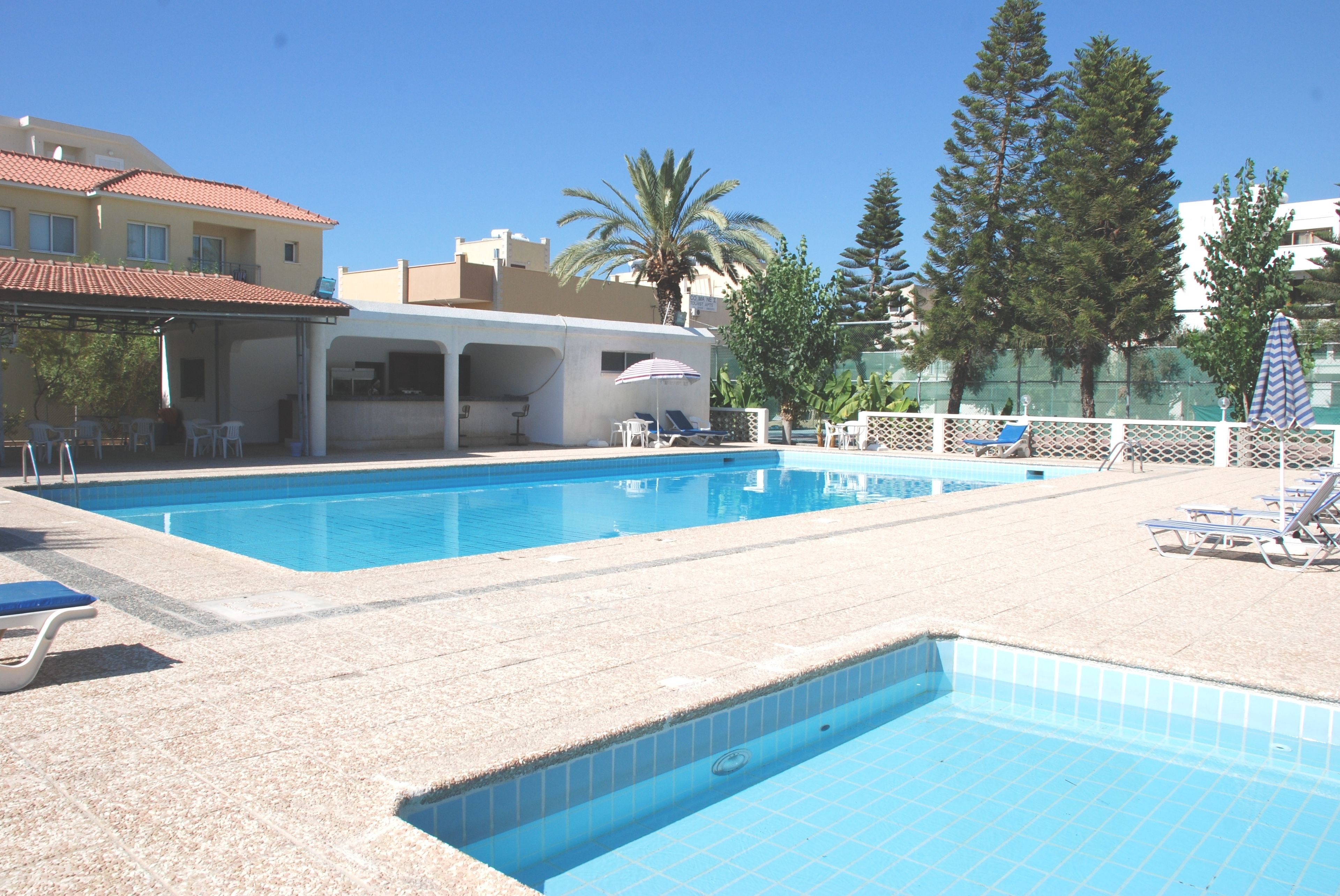 A swimming pool with lounge chairs and umbrellas, surrounded by buildings and trees.