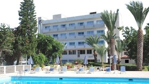 A swimming pool with lounge chairs and umbrellas, surrounded by palm trees and a multi-story building. - Marion (Polis)