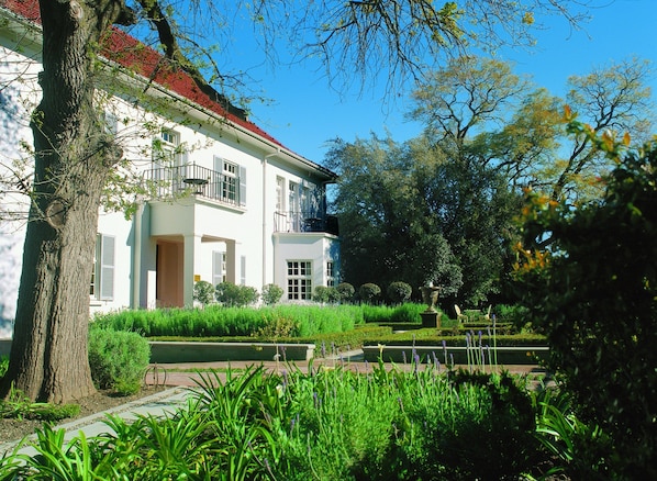 Un edificio blanco de dos pisos con balcón, rodeado de exuberante vegetación y jardines cuidados.