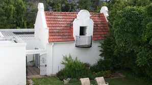 A white building with a red-tiled roof, a small balcony with a black railing, and two lounge chairs in the front yard. - Beauclair Guest Cottage (Stellenbosch)