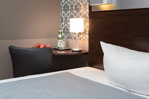 Una habitación de hotel con una cama bien tendida, una mesita de noche con una botella y vasos, y una silla.