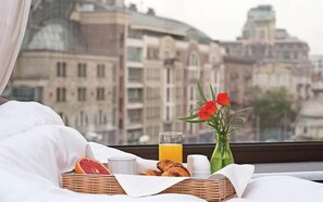 Ein Frühstücksangebot mit einem Korbwicker-Korb, der eine Grapefruit, Croissants und ein Glas Orangensaft enthält, einer Vase mit roten Blumen und einem weiteren Glas Orangensaft auf einem Bett mit Blick auf die Stadt.