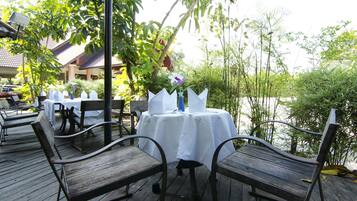 Un espacio de comedor al aire libre con mesas y sillas de madera, un mantel blanco y un florero con una flor.
