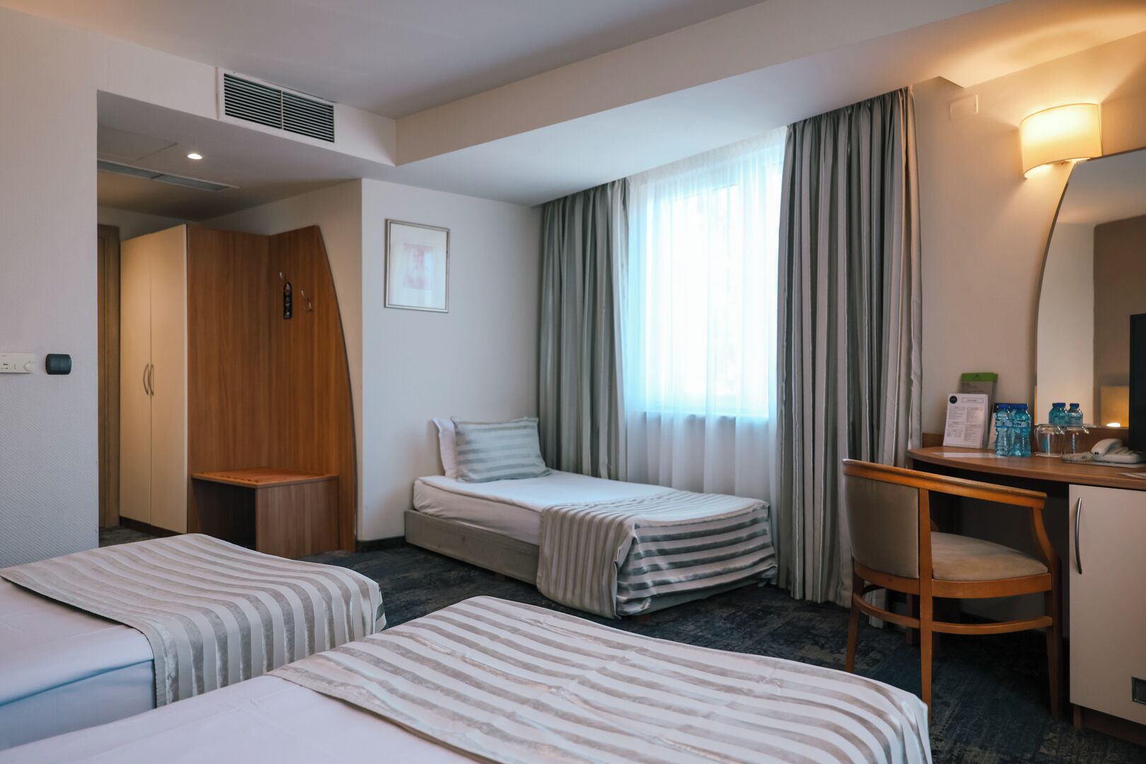 A hotel room with two single beds, a desk with a chair, a wardrobe, and a large window with curtains.