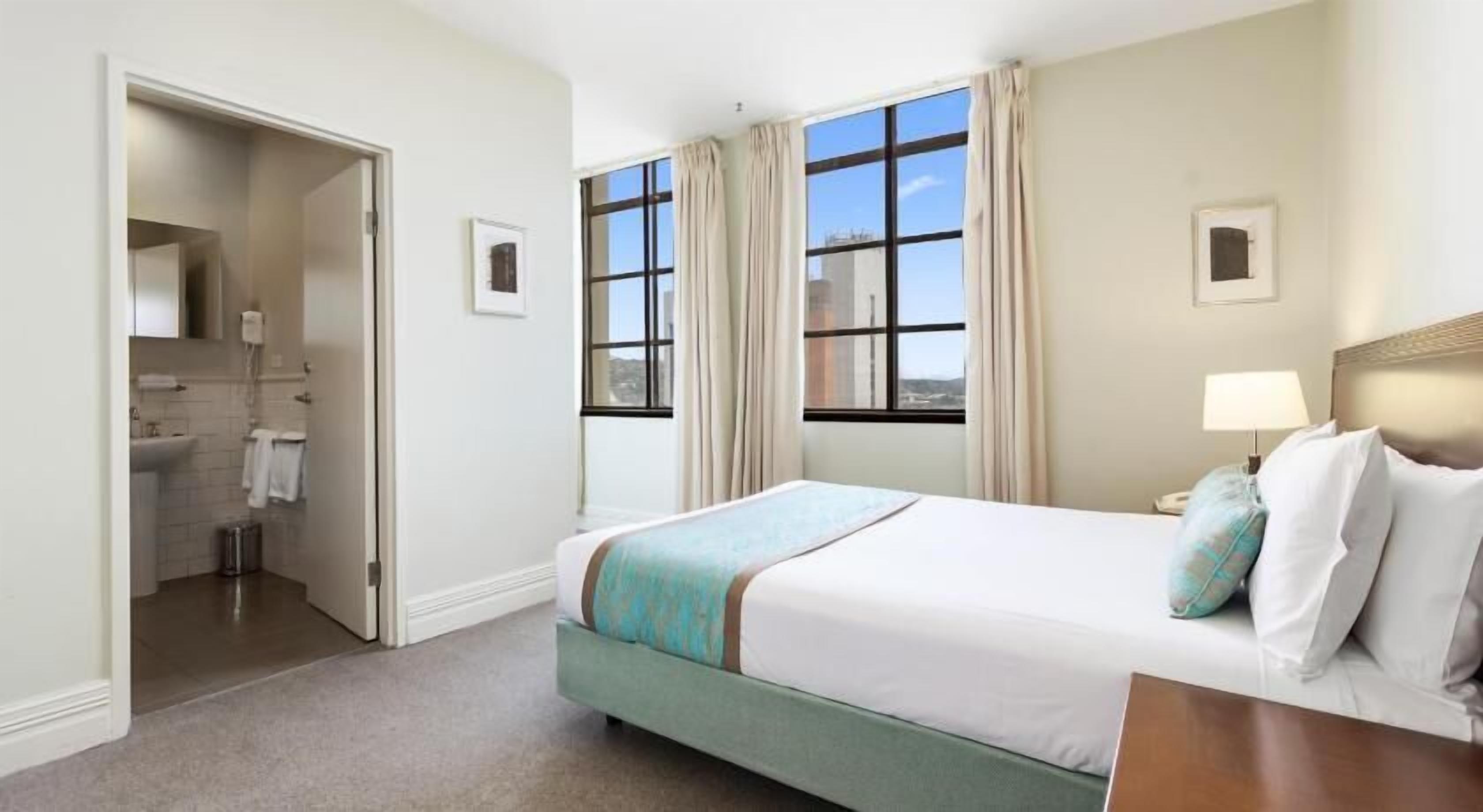 Una habitación de hotel con cama, mesita de noche, lámpara, ventana con cortinas, baño con lavamanos y toallero, y una puerta que da a otra habitación.