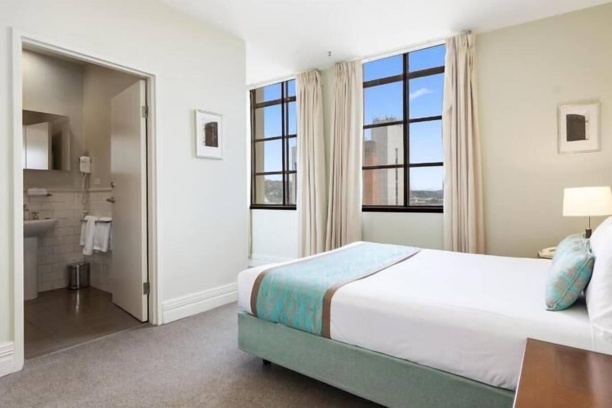 Una habitación de hotel con cama, mesita de noche, lámpara, ventana con cortinas, baño con lavamanos y toallero, y una puerta que da a otra habitación.