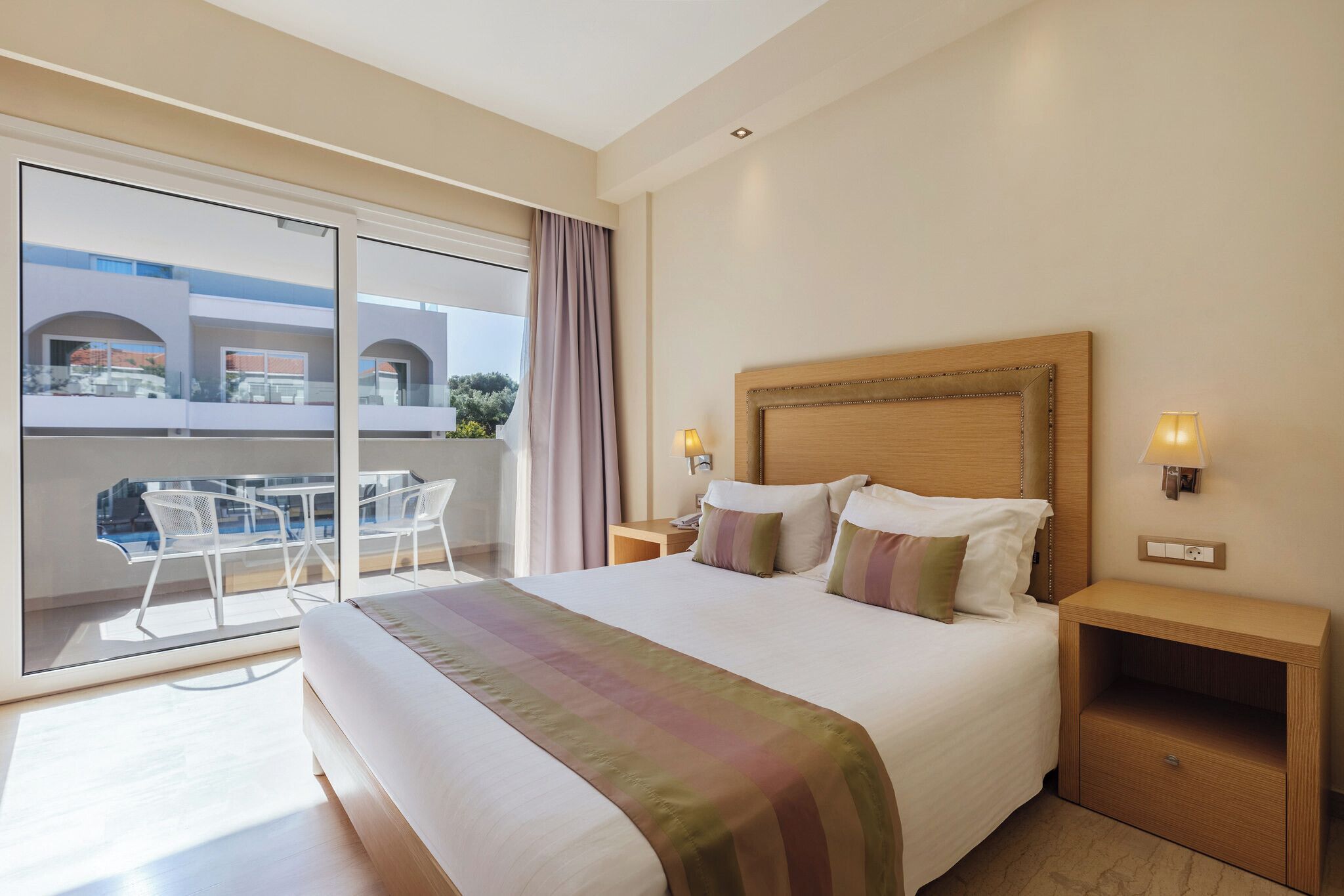 Habitación de hotel con una cama grande, una mesita de noche, una lámpara, un ventanal corredero a un balcón con mobiliario exterior y vistas a edificios.