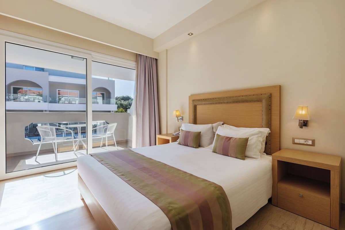 Habitación de hotel con una cama grande, una mesita de noche, una lámpara, un ventanal corredero a un balcón con mobiliario exterior y vistas a edificios.