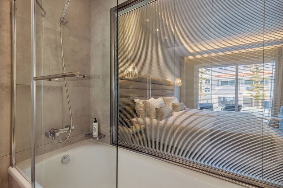 A modern bathroom with a glass shower enclosure, a bathtub, and a view of a bedroom with a bed and a window.