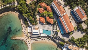 An aerial view of a coastal resort with a swimming pool, beach, and multiple buildings with red-tiled roofs.