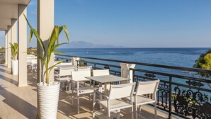 A terrace with white chairs and tables, potted plants, and a view of the sea.