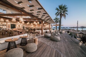 Un área de comedor al aire libre, moderna, con mesas y sillas de madera, luces colgantes y vista a la playa.