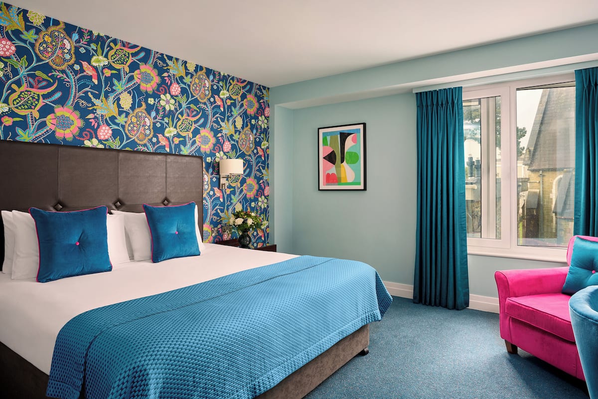 Quarto de hotel com uma cama grande, um sofá rosa, uma janela com cortinas e uma parede com padrão floral.