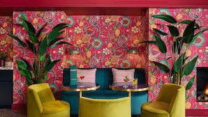 Una habitación vibrante con un sofá azul turquesa, sillas amarillas y un llamativo papel tapiz floral.