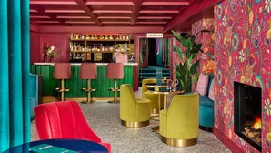 Zona de bar con mostrador de recepción, taburetes y una variedad de botellas. Hay sillas y una mesa con un florero.