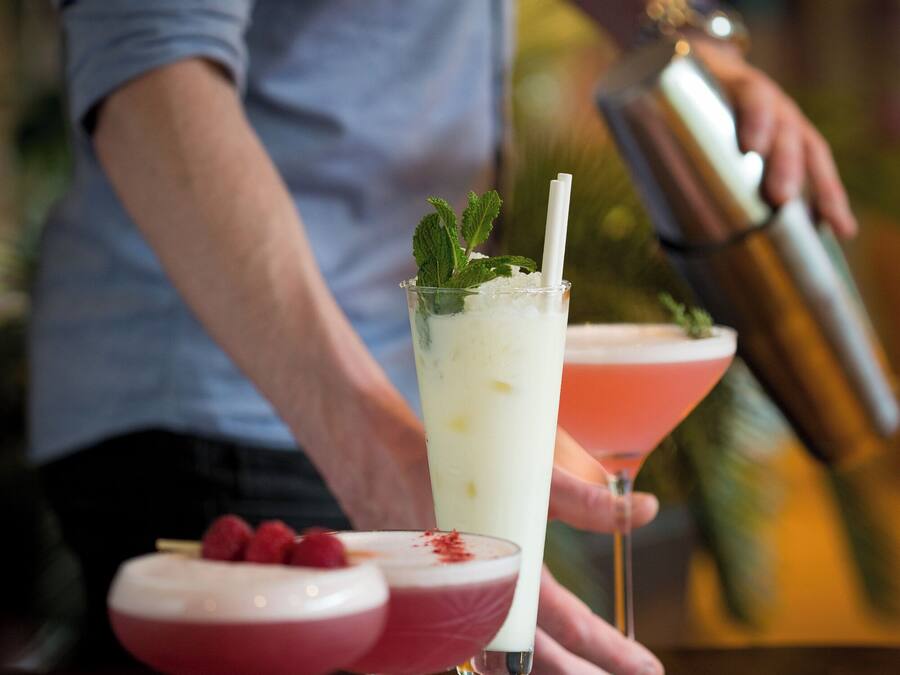 Un bar con una variedad de bebidas, incluyendo un vaso alto con pajita y adornado con hojas de menta, y un cóctel rosado con frambuesas.