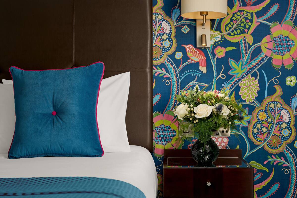 Uma cama com travesseiro azul e lençóis brancos, parede com padrão floral e um vaso de flores em uma mesa de cabeceira.