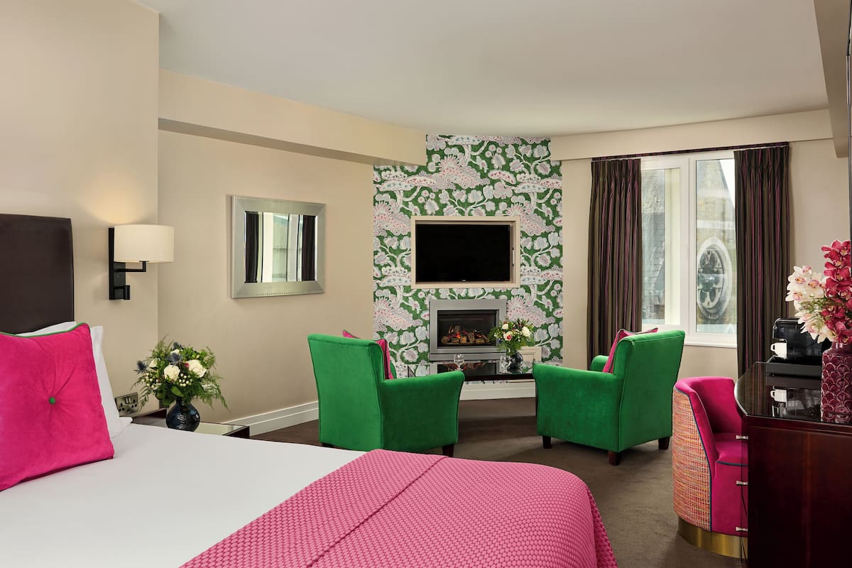 Quarto de hotel com cama, duas poltronas verdes, lareira, televisão na parede e janela com cortinas.
