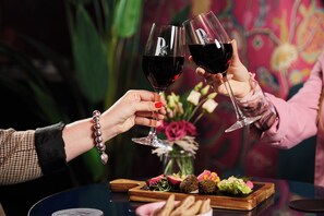 Two people toasting with wine glasses, a wooden board with food, and a vase with flowers.