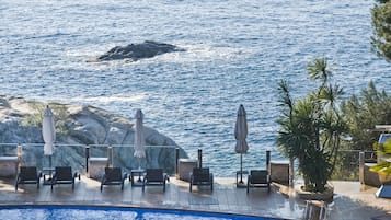 Un área junto a la piscina con sillones de descanso y sombrillas, con vistas a un cuerpo de agua donde se divisa una ballena a lo lejos.