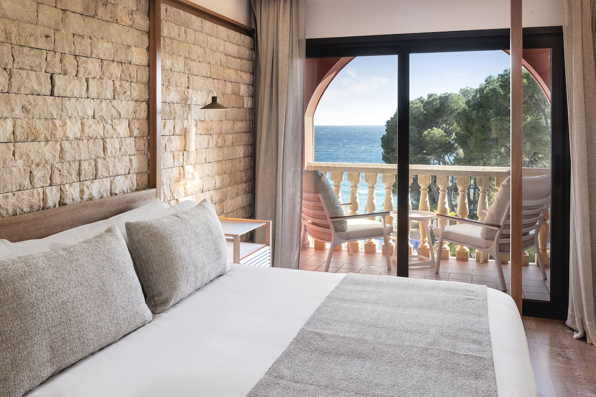 Une chambre avec un grand lit, une vue sur la mer depuis un balcon ouvert et un mur en briques.