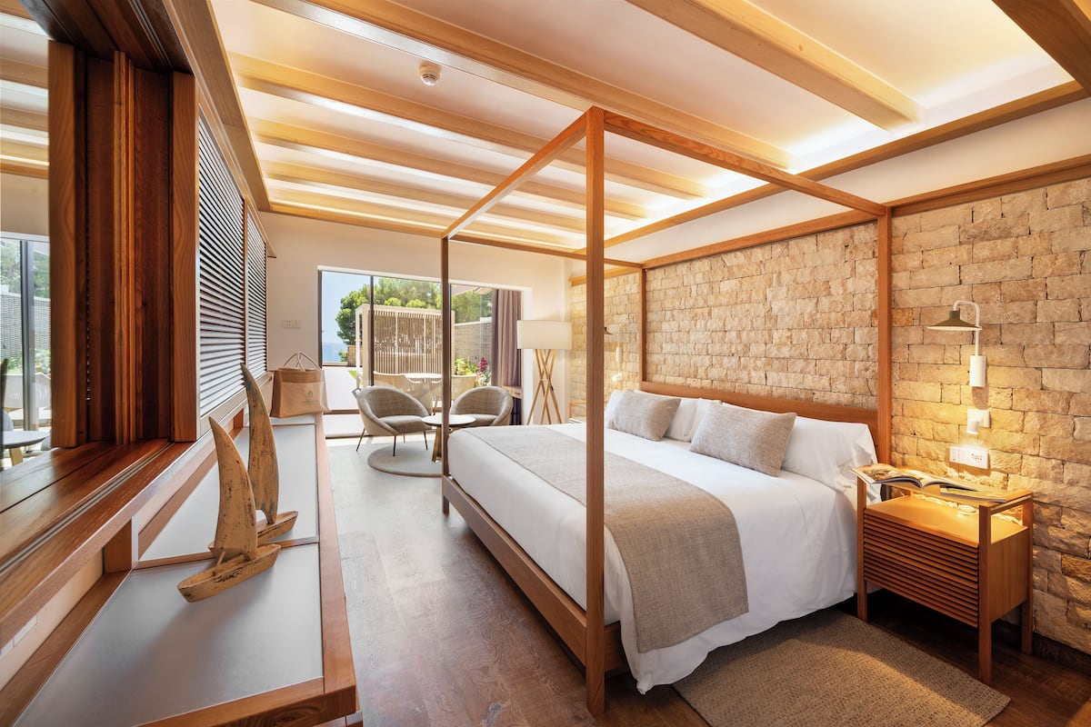 Une chambre moderne avec un lit à baldaquin, des poutres en bois, un mur en pierre et une vue sur l’extérieur.