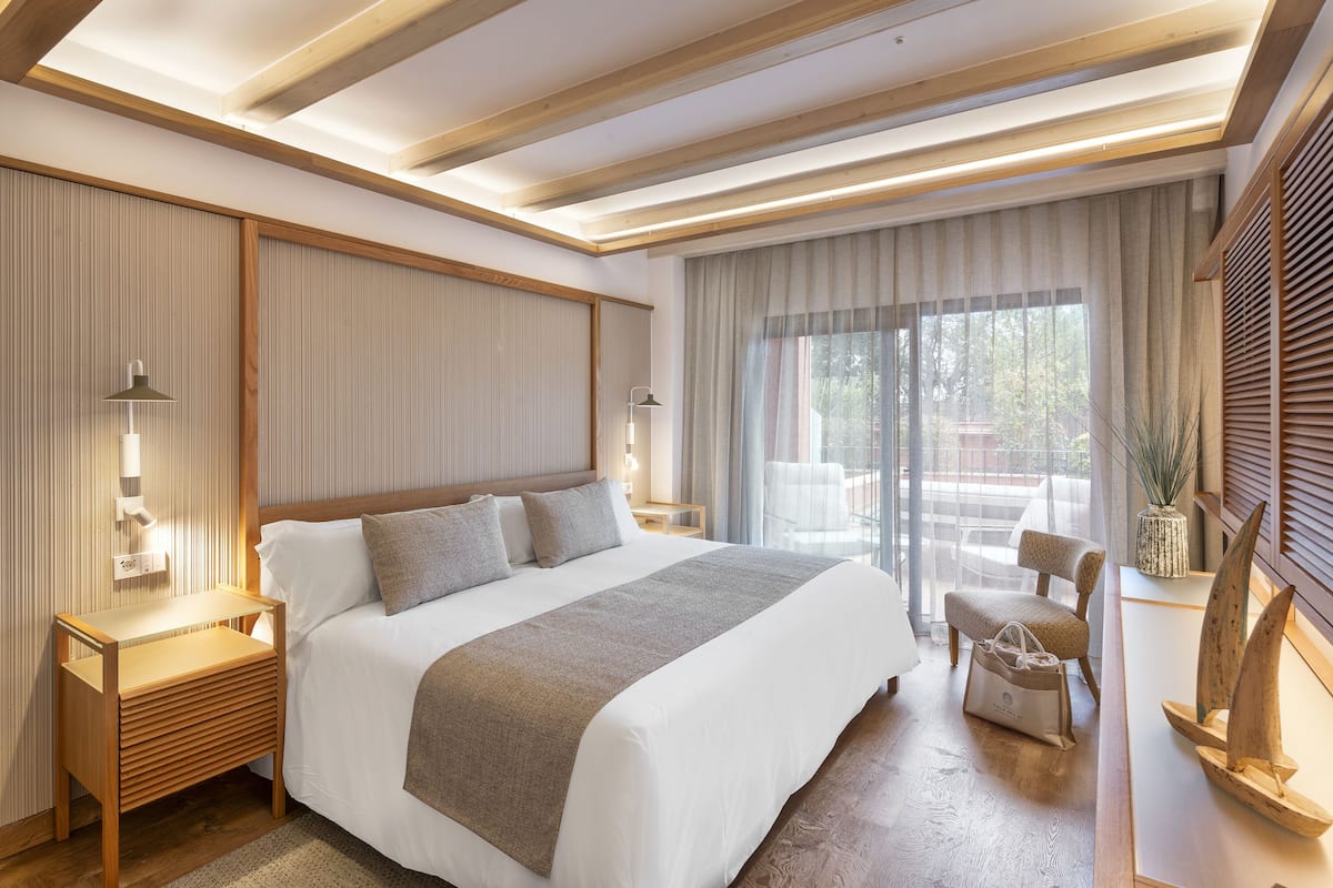 Une chambre d’hôtel moderne avec un grand lit, une tête de lit en bois, des tables de chevet avec des lampes, un coin salon avec un fauteuil et une vue par la fenêtre.