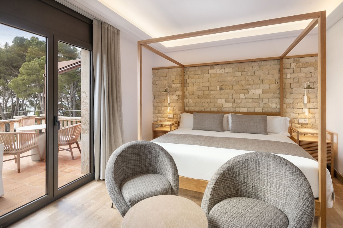 Une chambre moderne avec un grand lit, deux tables de chevet, une tête de lit ornée d’un motif en briques et un balcon avec un espace salon extérieur.