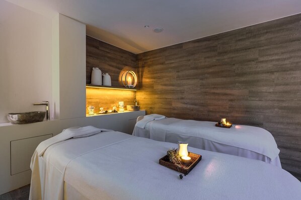 A modern spa room with two beds, a wooden wall, a sink with a vessel basin, and a shelf with candles and decorative items.