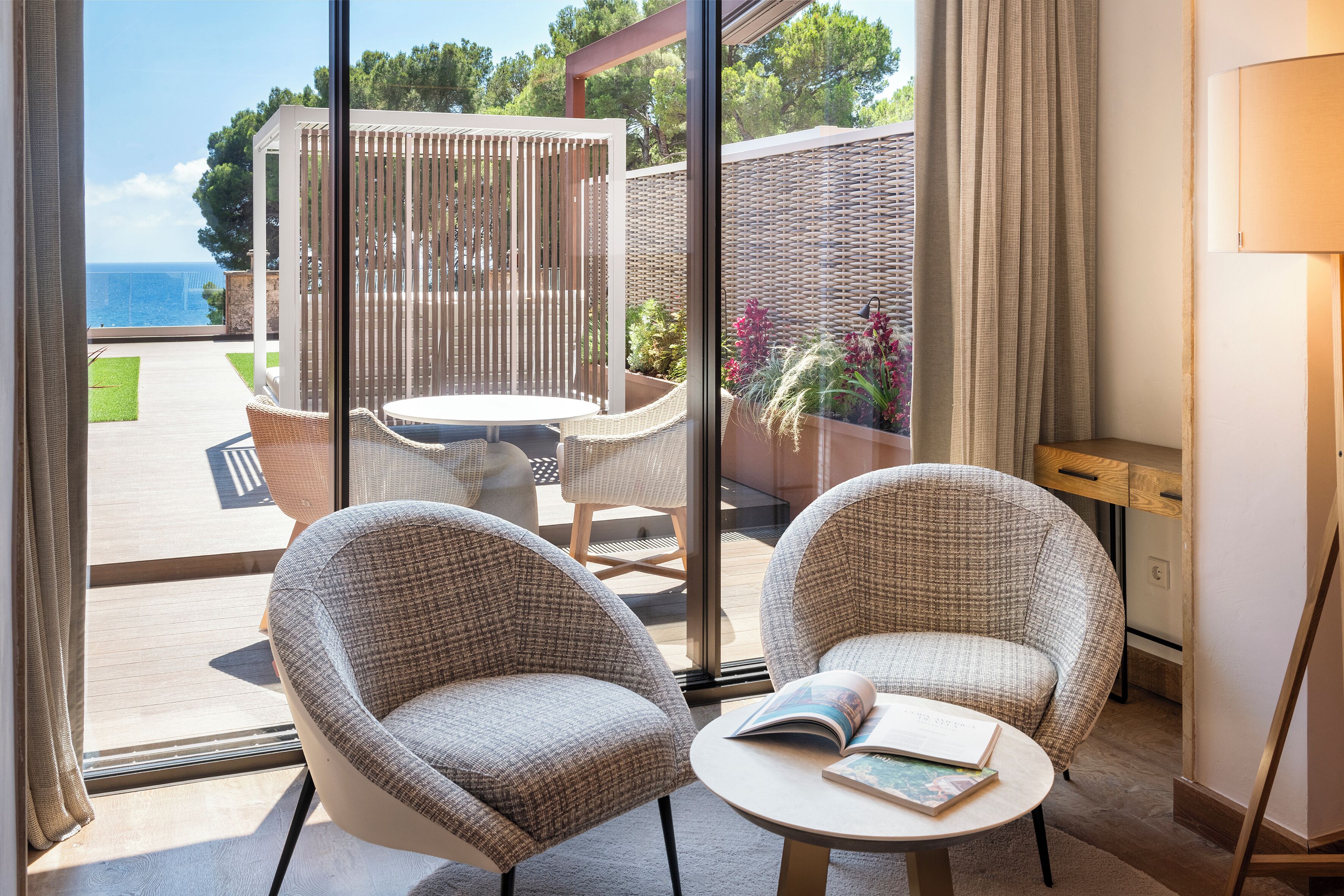 A room with two wicker chairs, a small table, and a view of the sea through large windows.
